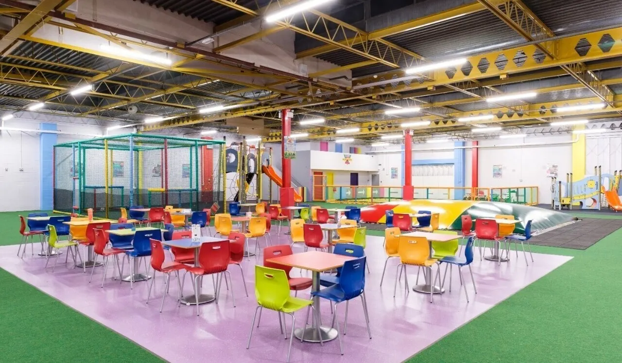 Colourful indoor play centre with tables, bright chairs, climbing equipment and slides under industrial yellow ceiling beams.