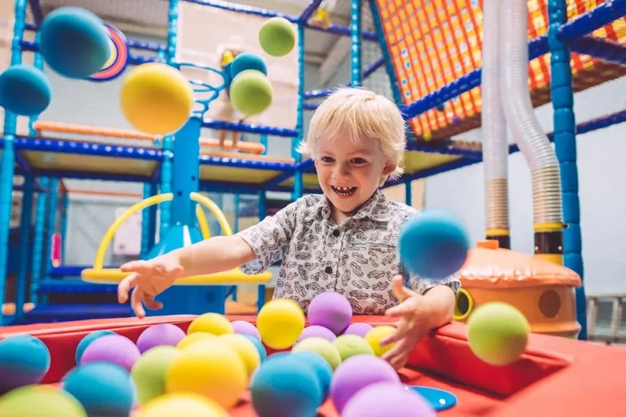 Young child playing with coloured balls in an indoor soft play area with blue climbing structures.