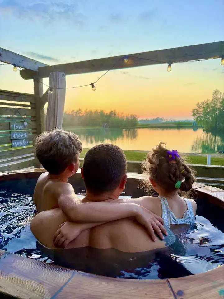 Three children in a hot tub at sunset overlooking a calm lake with woodland beyond.