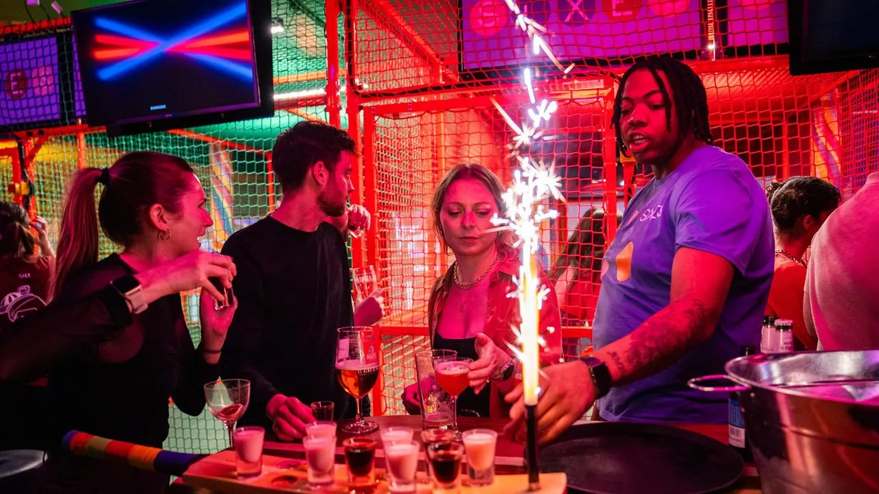 Group of people enjoying drinks and socialising in a vibrant neon-lit venue with red mesh cages and glowing signage.