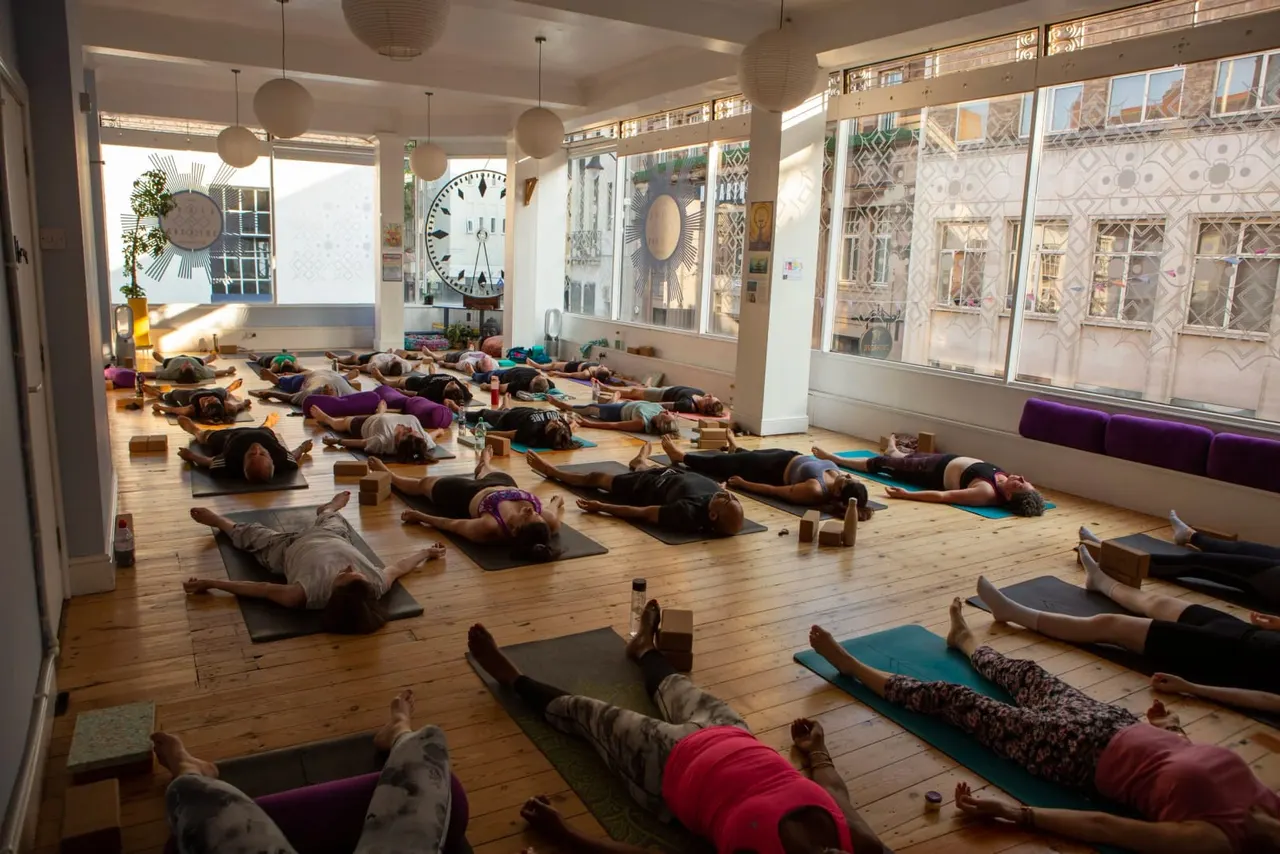 Large group of people lying on yoga mats in a bright, airy studio during a relaxation or yoga class.