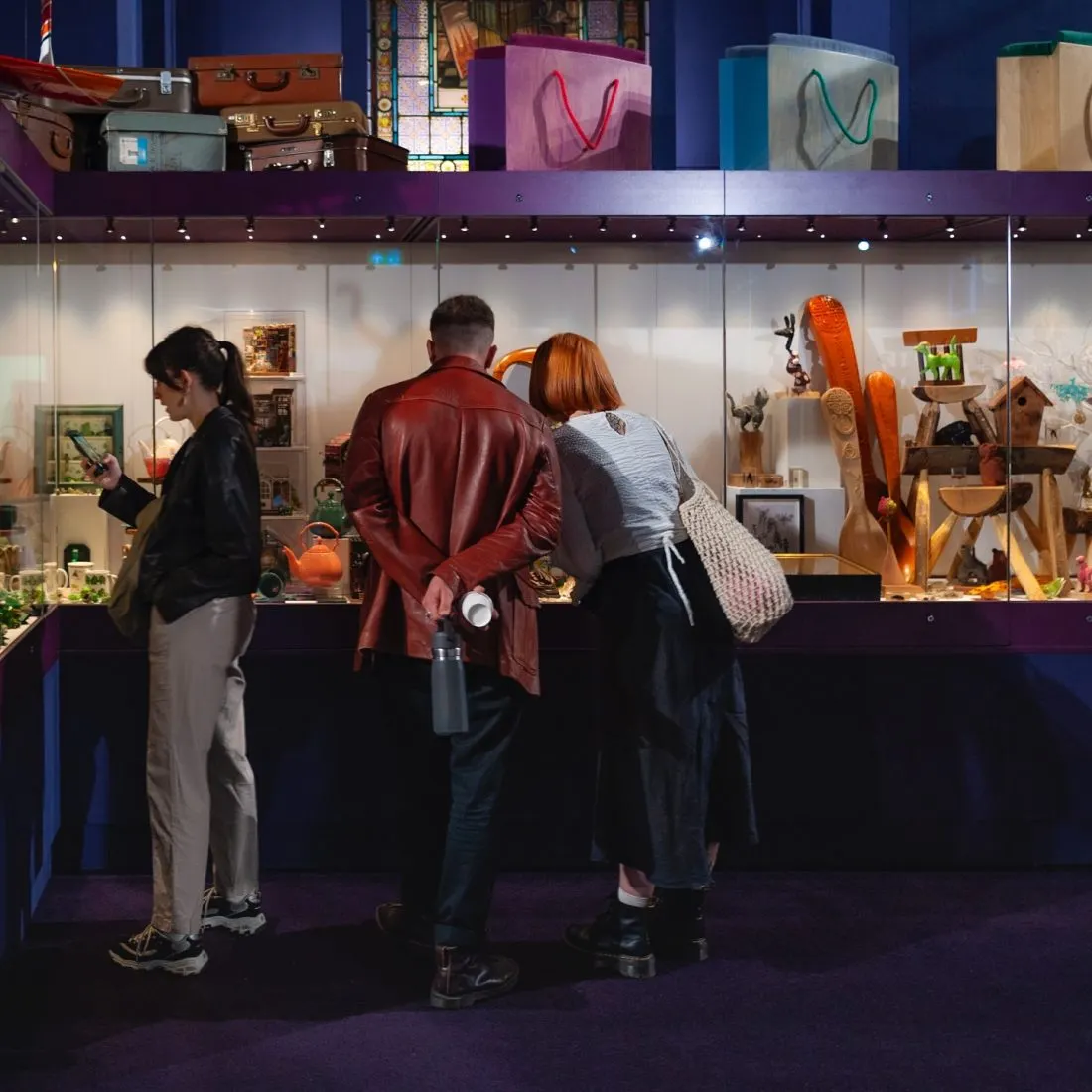 Three visitors viewing a glass display case containing wooden carved items and vintage objects in a museum gallery.