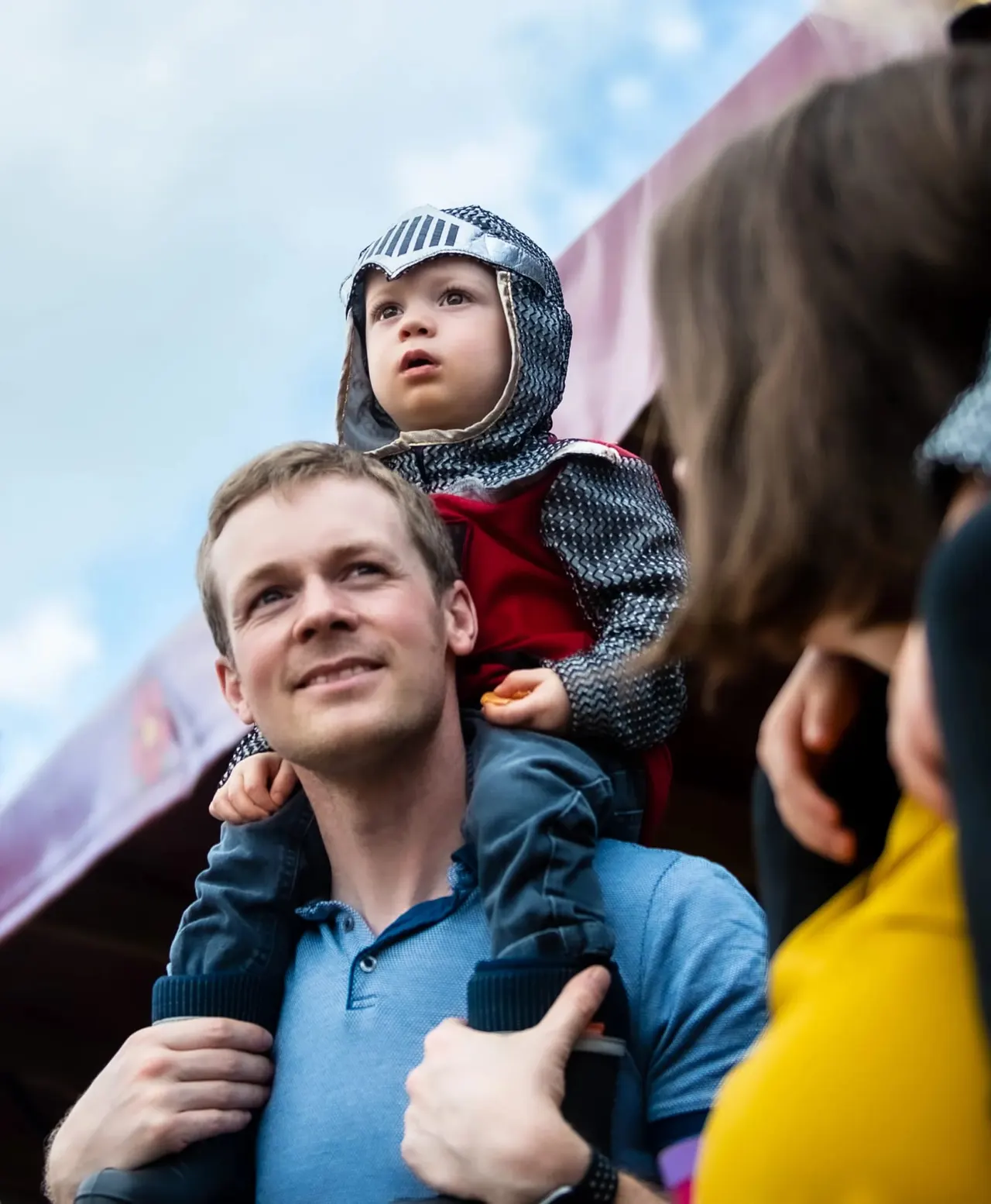 A young child in medieval chainmail and helmet sits on a man's shoulders whilst others watch at an outdoor venue.