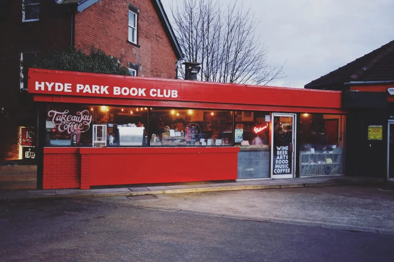 Bright red storefront of Hyde Park Book Club café with takeaway coffee sign and illuminated windows, exterior view at dusk.