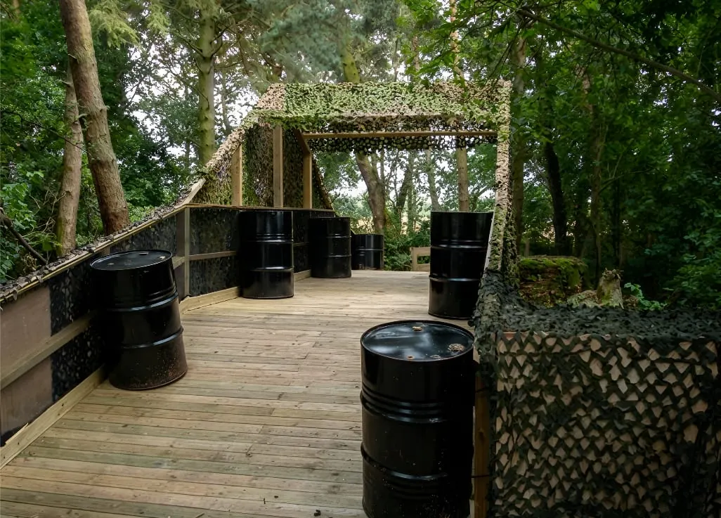 Woodland paintball arena with black barrel bunkers and camouflaged pergola shelter on wooden deck.