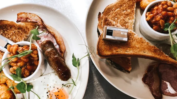 Two white plates with grilled sausage, buttered toast, baked beans and microgreens garnish on a table.