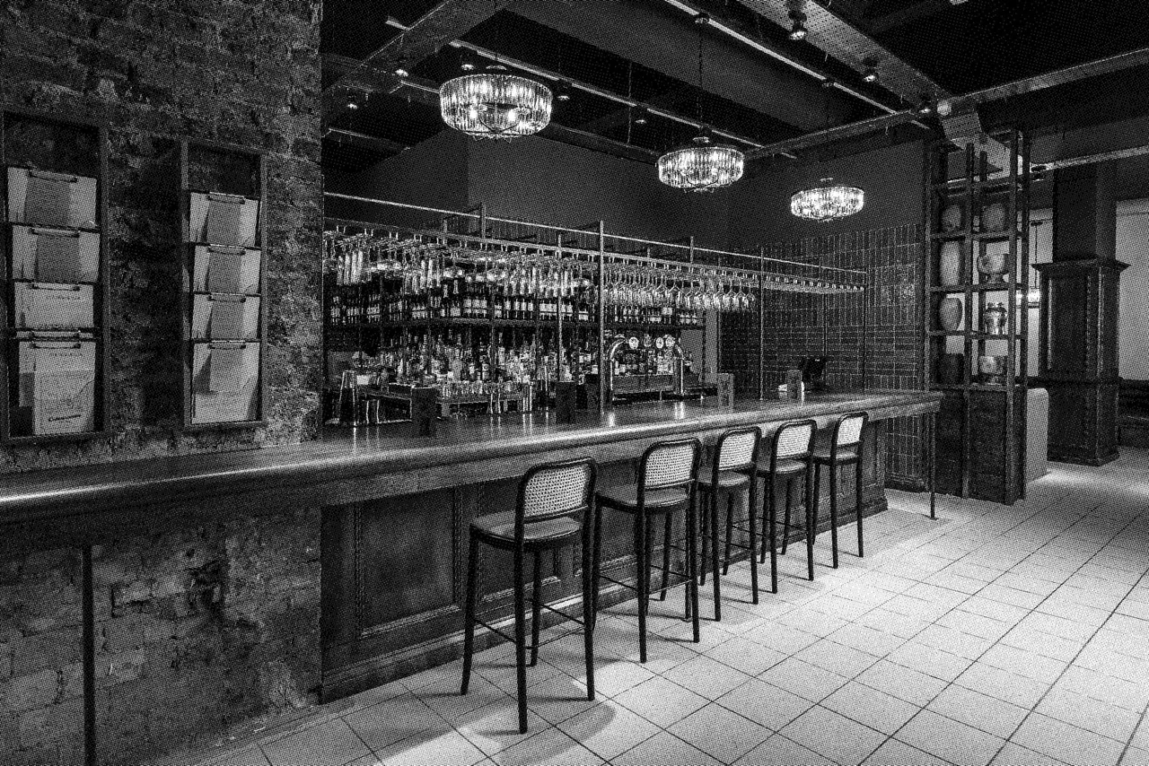 Modern bar interior with long counter, bar stools, shelves of bottles, pendant lighting and exposed brick pillars.
