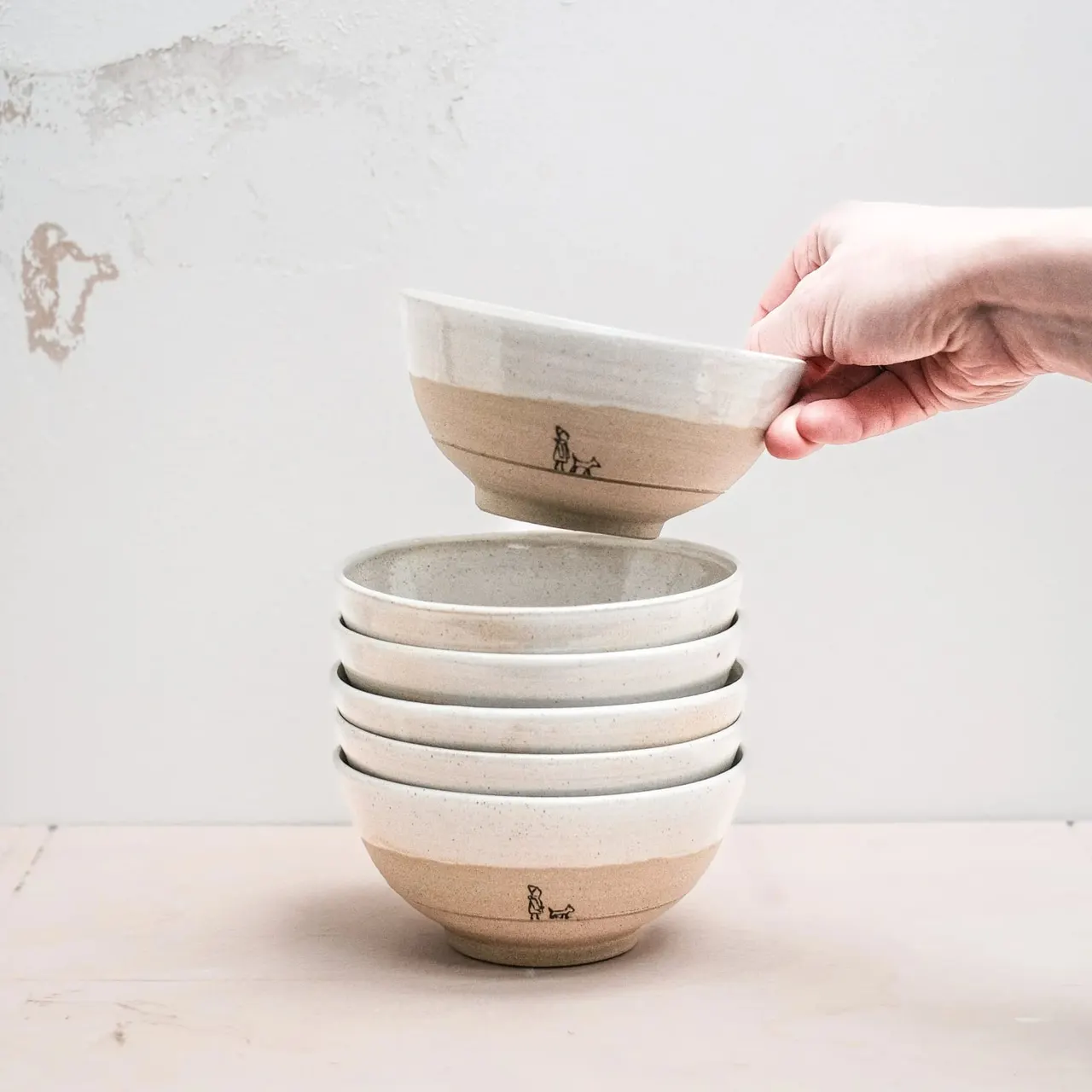 Hand placing a ceramic bowl onto a stacked collection of matching neutral-toned bowls with maker's mark.