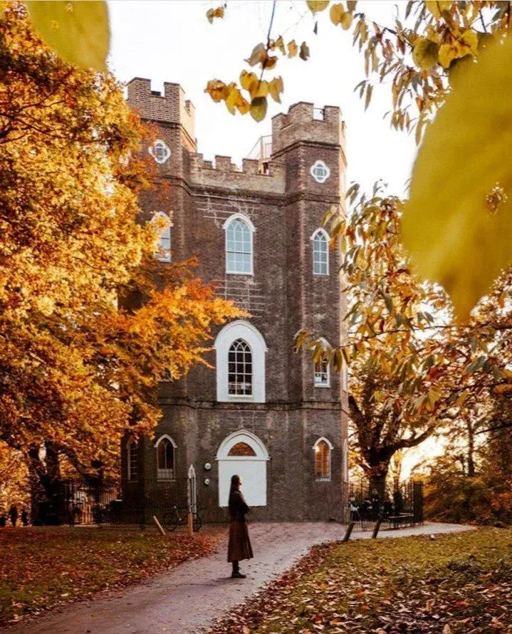 A person stands before a brick castle tower with crenellations, surrounded by autumn foliage in golden tones.