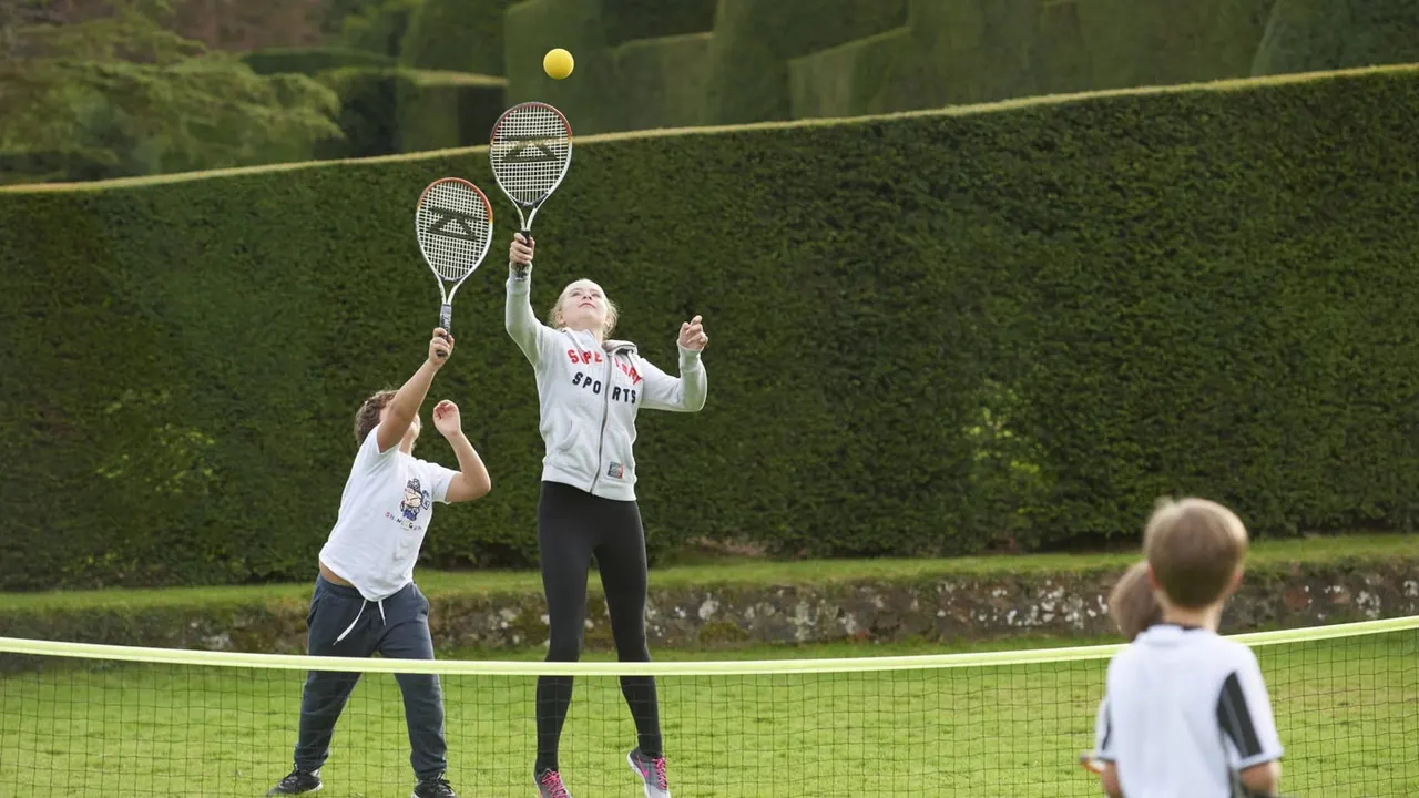 Young children playing tennis on a grass court bordered by tall clipped hedges.