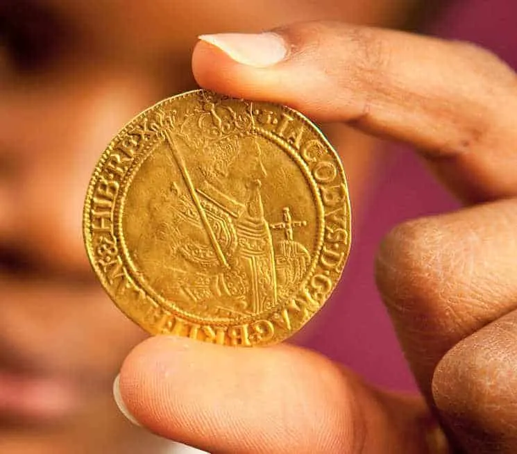 Close-up of a person's hand holding an antique gold coin with ornate detailing against a blurred background.
