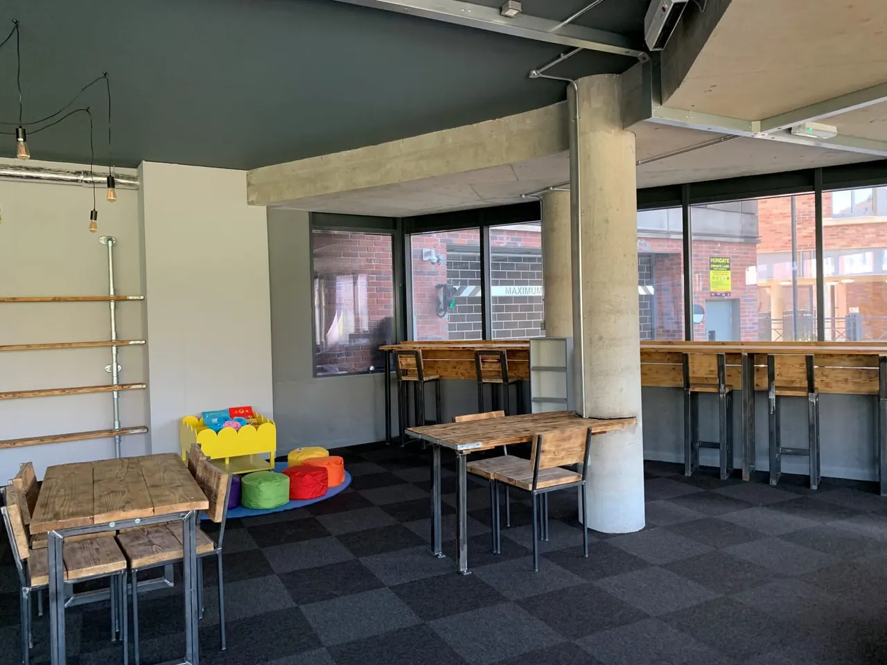 Modern café interior with wooden tables, chairs, colourful soft seating, and large windows overlooking brick buildings.