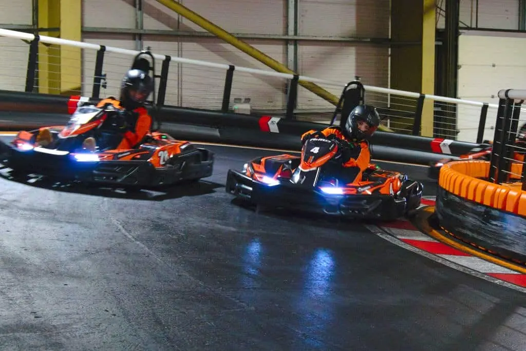 Three people in helmets racing orange go-karts with blue lights around an indoor track with barriers.