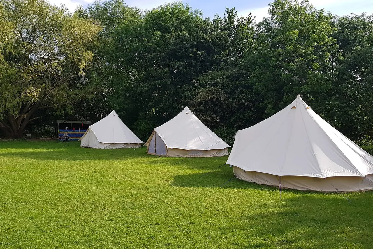 Three cream bell tents pitched on a grassy meadow surrounded by mature trees.