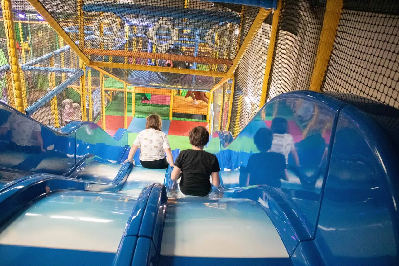 Two young children sitting in a blue slide at an indoor play centre with coloured foam pads and netting visible above.