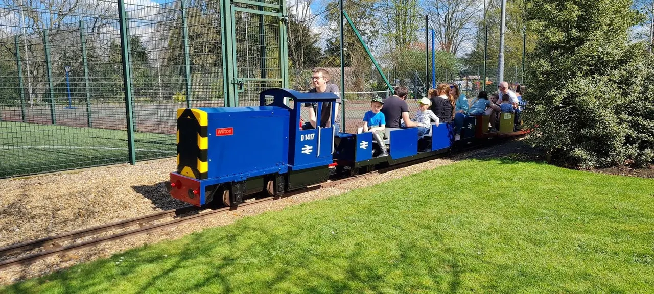 A blue miniature steam train with passengers riding in open carriages along a narrow-gauge track in a park setting.