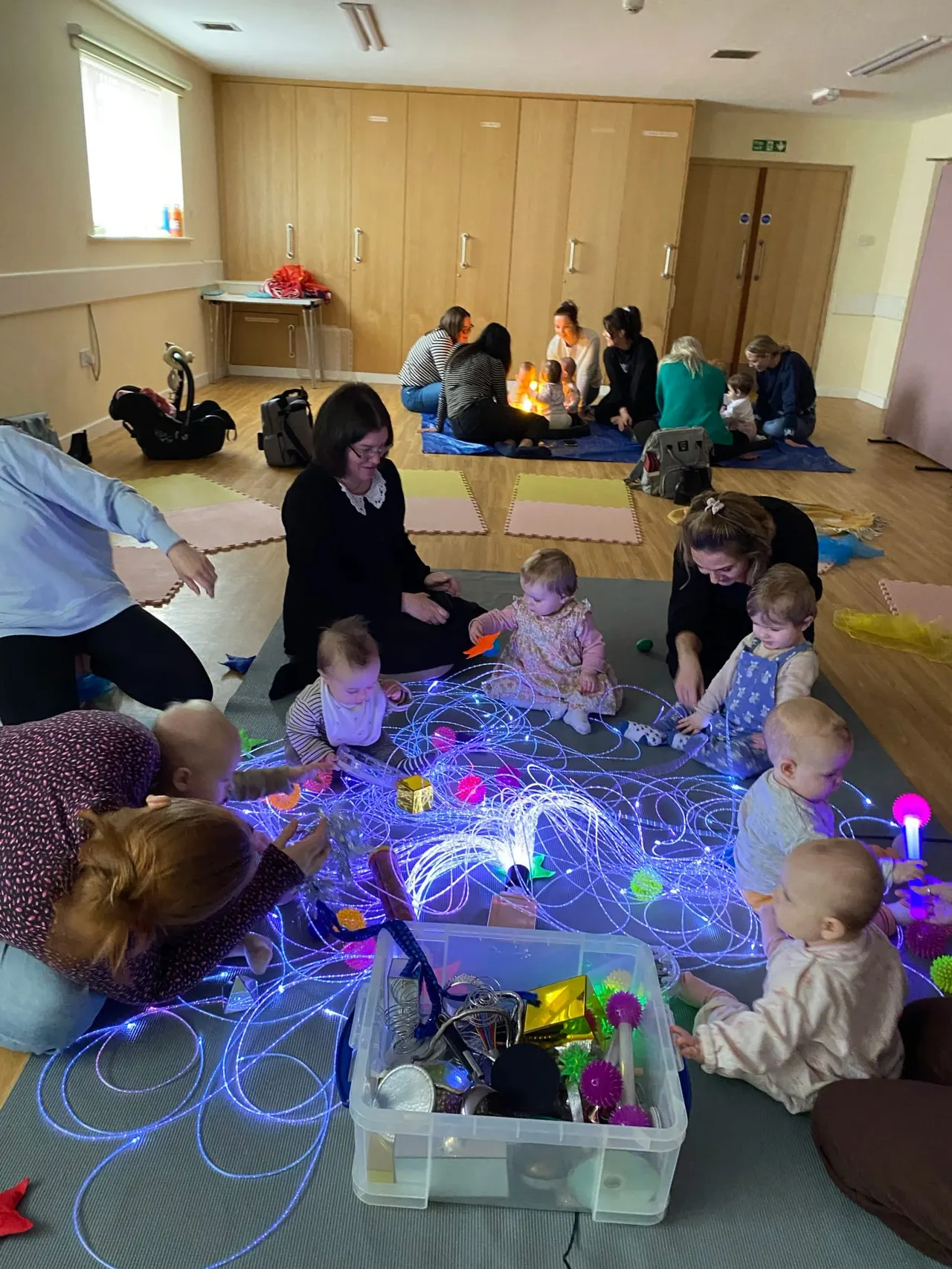 Toddlers and adults engage with glowing interactive light panels and sensory toys on the floor of an indoor play space.
