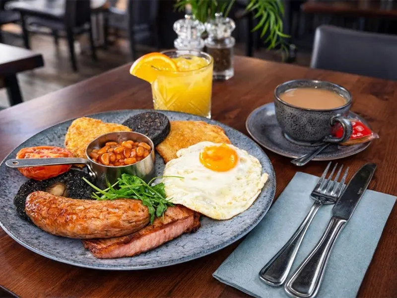 Full breakfast plate with sausage, bacon, fried egg, beans and toast, served with orange juice and coffee.
