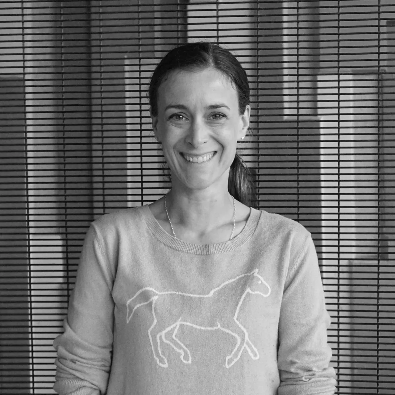 A smiling woman wearing a horse-print jumper poses against a slatted wall background.