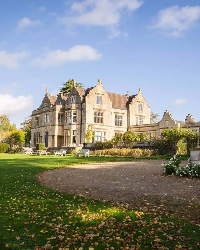 Victorian stone mansion with manicured grounds and driveway under blue sky with white clouds.