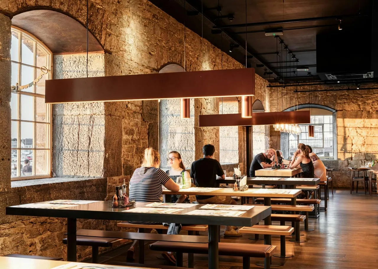 Interior of an industrial-style venue with exposed stone walls, wooden tables and benches, and several visitors seated and dining.