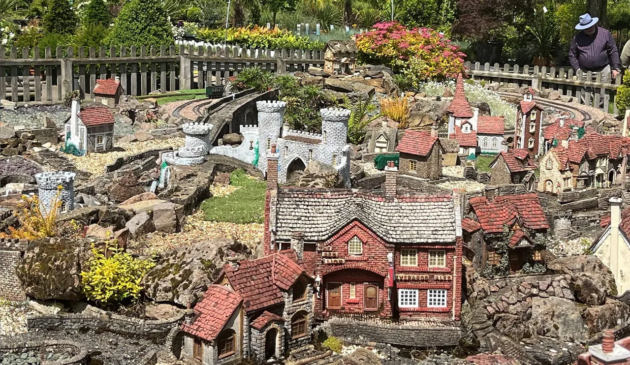 Miniature model village with detailed buildings, castle towers, and landscaped grounds featuring colourful flowers and rock formations, with visitors viewing from above.