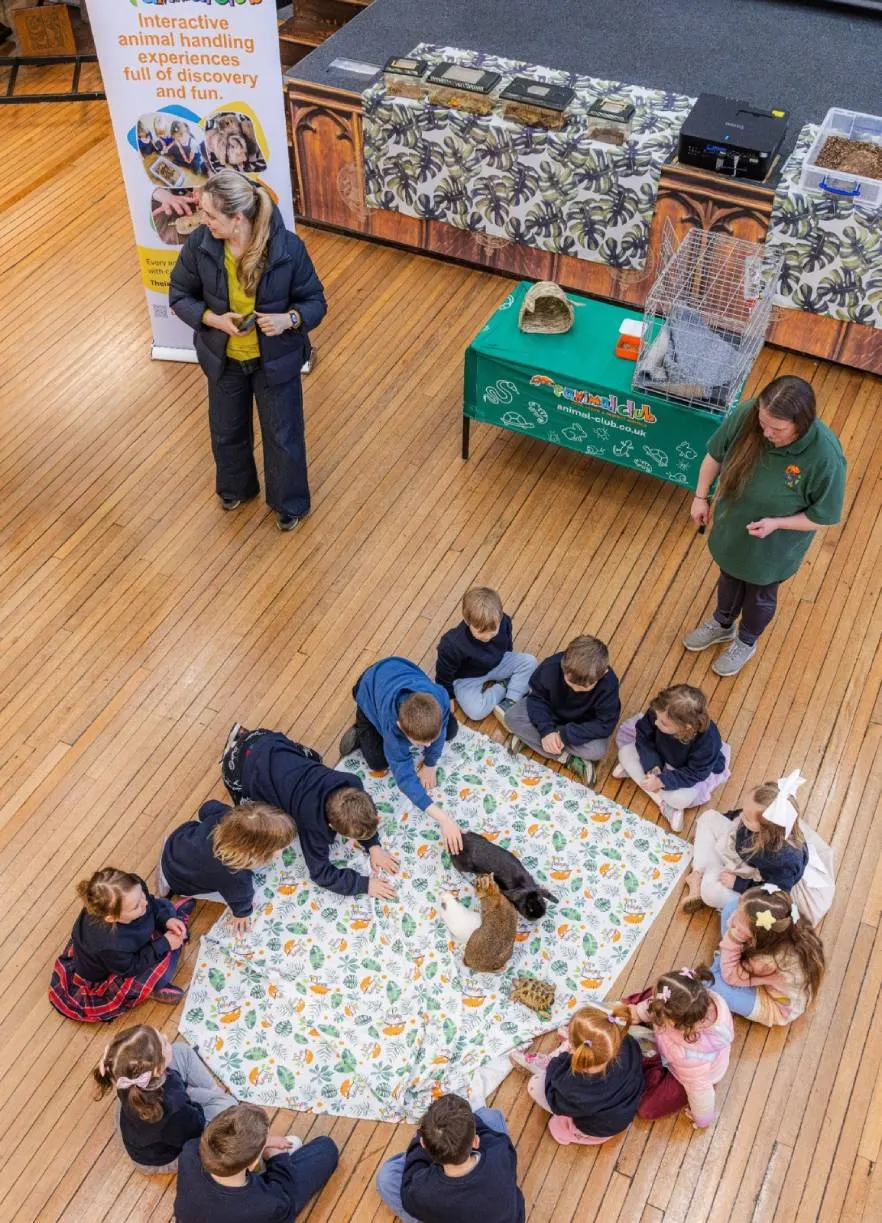 Children sit in a circle on a patterned cloth with farm animals during an interactive animal handling experience session indoors.