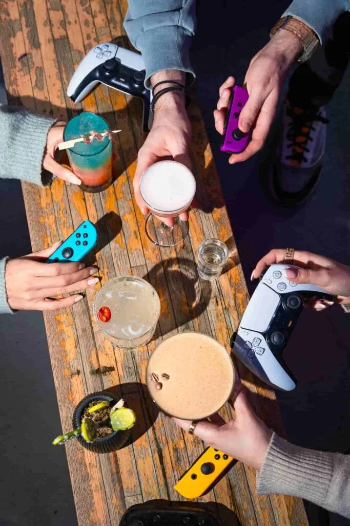 Group of people holding gaming controllers and drinks around a wooden table.