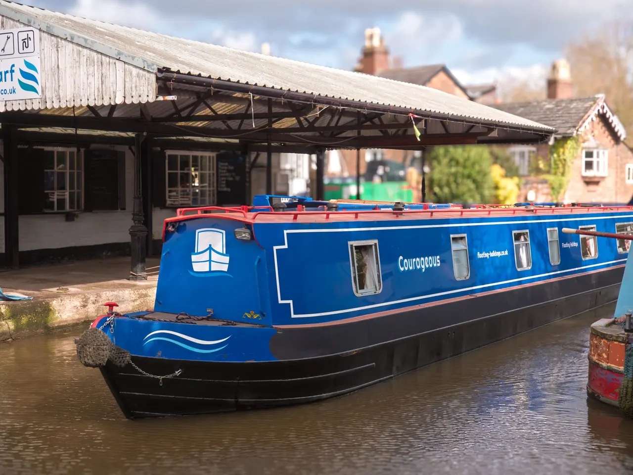 Blue and black narrowboat moored at a canal-side facility with brick buildings and covered structures behind.
