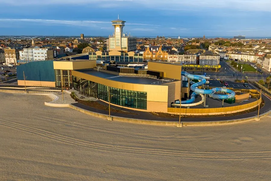 Aerial view of a modern yellow leisure centre with water slides on a beach, overlooking a coastal town.