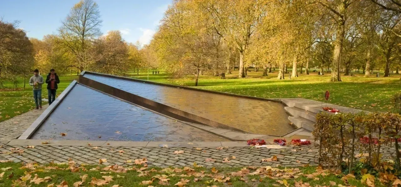 A modern water feature in an autumn park with visitors walking nearby, surrounded by golden trees and fallen leaves.