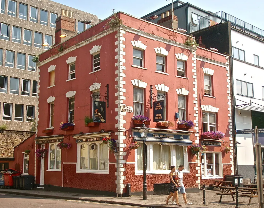 Historic red-brick pub building with white stone detailing and hanging flower baskets, pedestrians walking past on street.