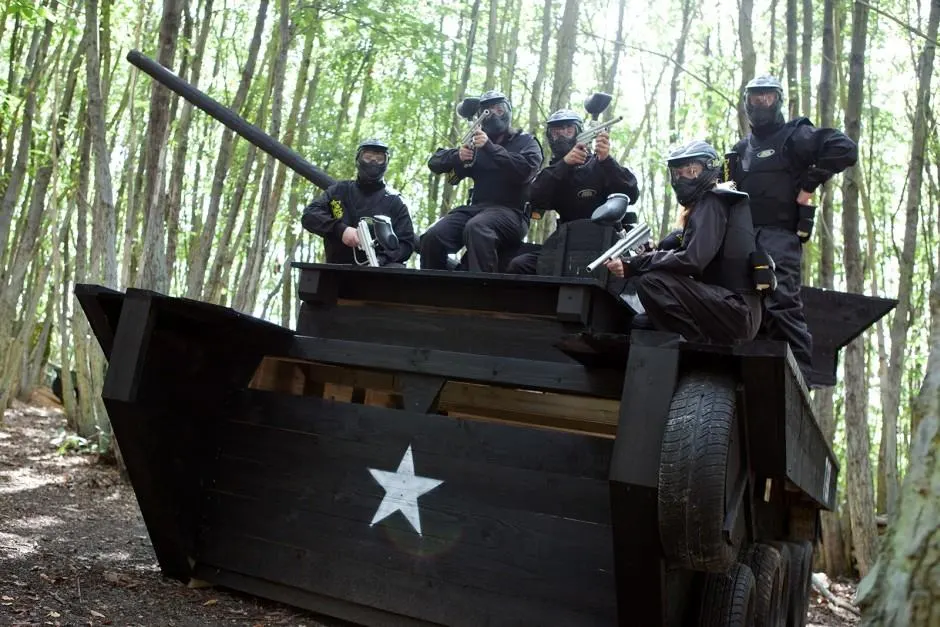 Group of players in protective gear and helmets posing on a black pirate-themed vehicle in a wooded paintball arena.
