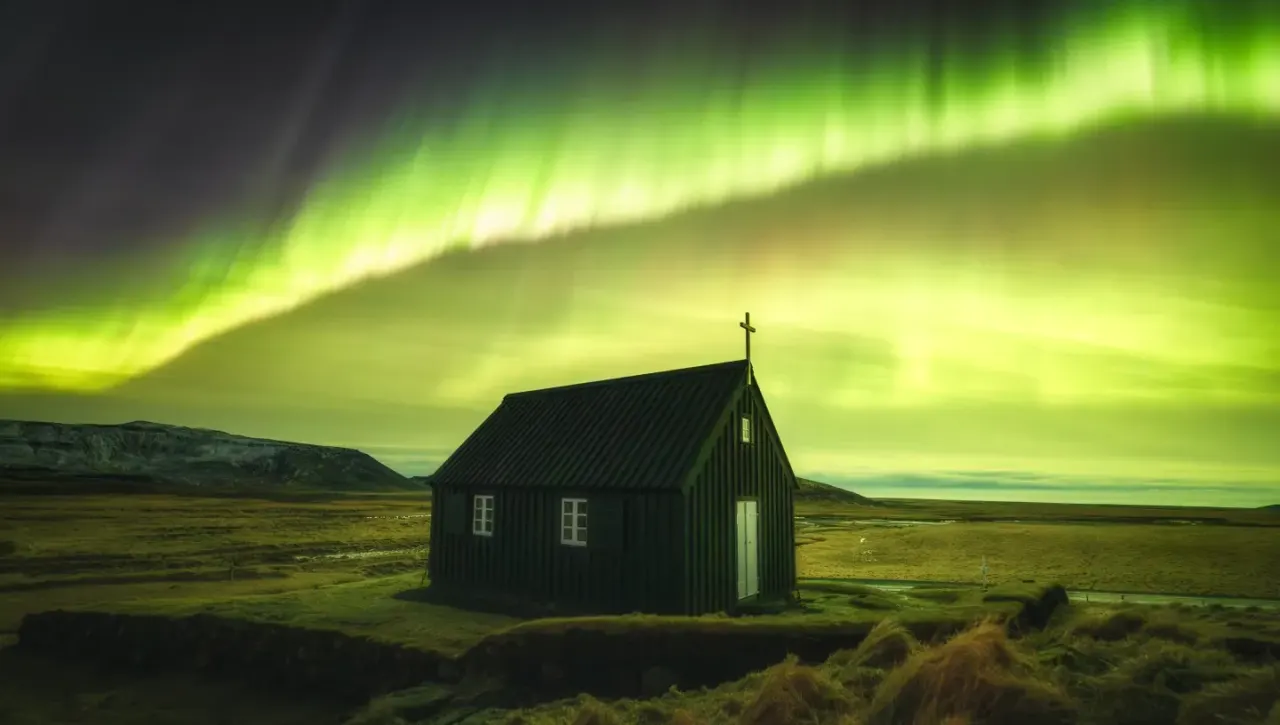 This is a photograph, not a designed graphic.

Small black corrugated-iron chapel with a cross on its roof, set in an Icelandic moorland landscape under a glowing green northern lights sky.