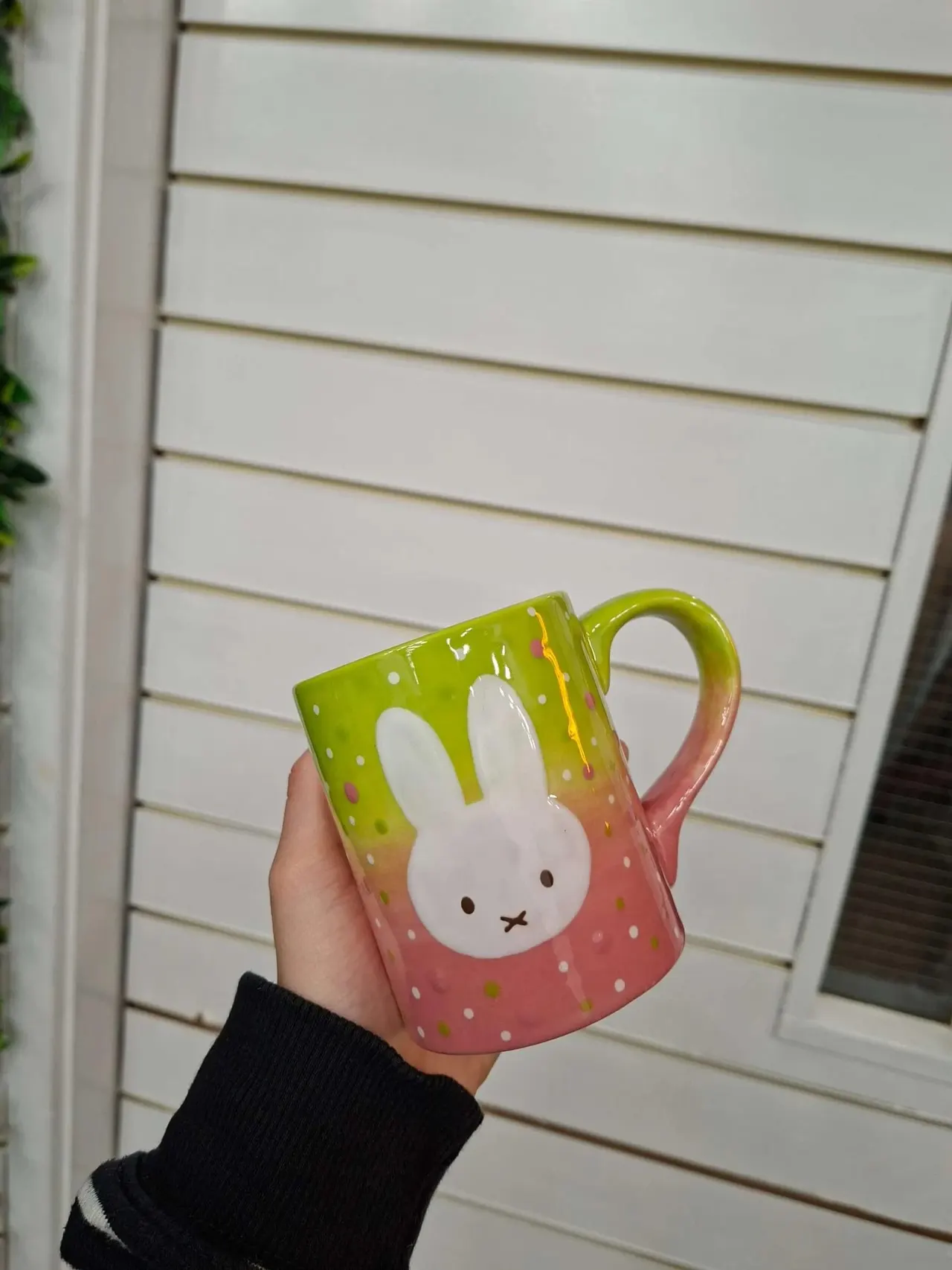 Hand holding a colourful ceramic mug decorated with a rabbit face design against a white wooden wall.