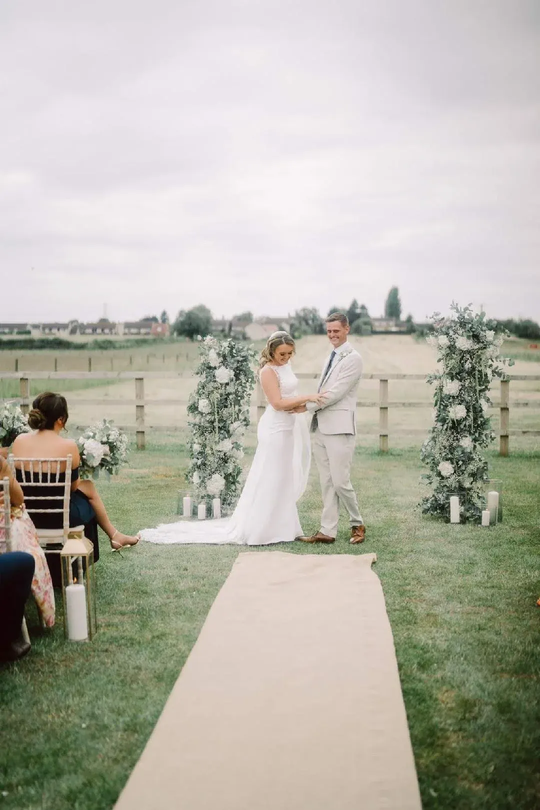 Bride and groom exchange vows at outdoor wedding ceremony in rural field with floral arch and guests seated alongside.