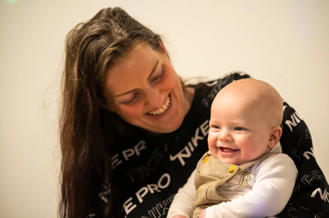 Woman holding a smiling baby during a music class session against a neutral background.