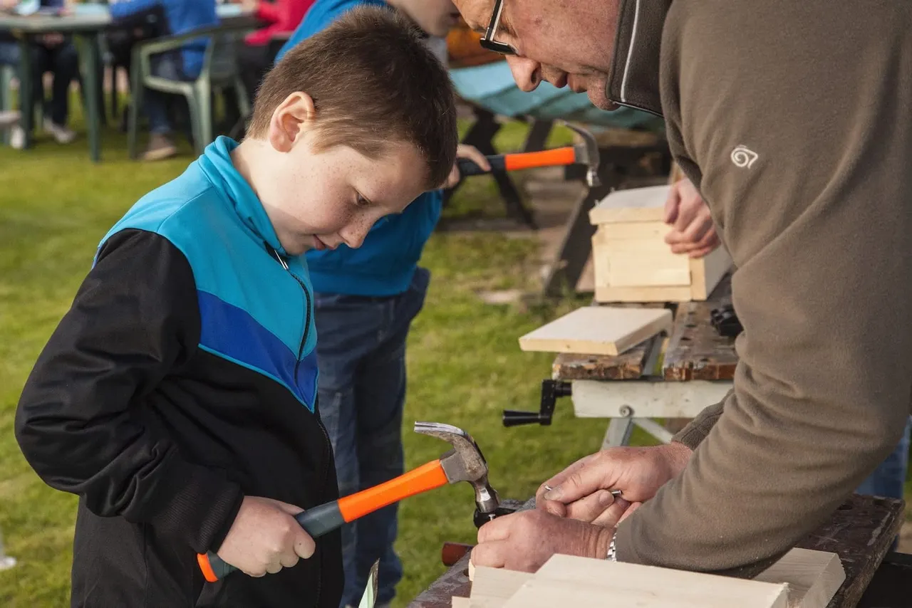 A young boy in a blue and black jacket learns woodworking from an adult instructor, holding a hammer at an outdoor craft activity on a grass area.