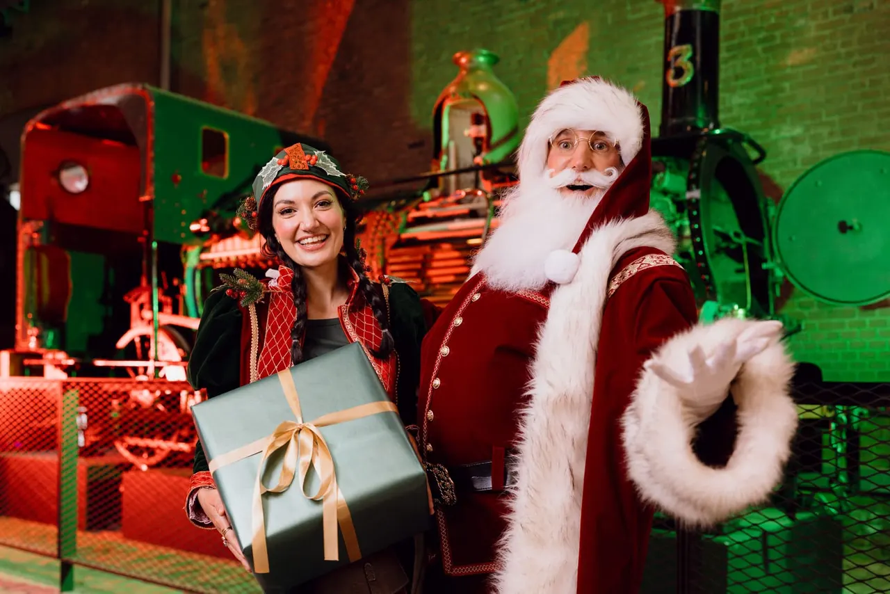 A woman and a man dressed as Father Christmas stand with a gift in front of a decorated toy train display.