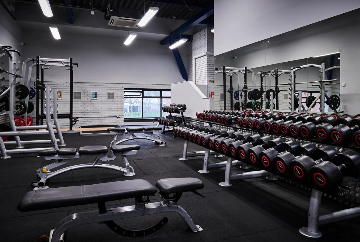 Modern gym interior with rows of red and black dumbbells, exercise benches, and weight training equipment.