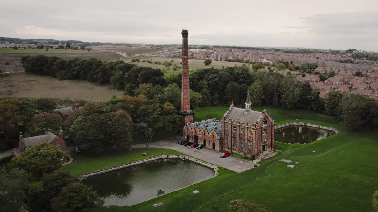Aerial view of a Victorian red-brick pumping station with a tall chimney beside a pond, surrounded by green fields and residential areas.