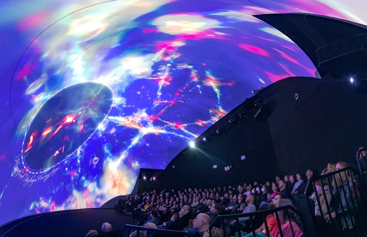 Visitors sit in a curved theatre watching a vibrant digital projection of Earth's surface with glowing city lights on a vast curved screen.