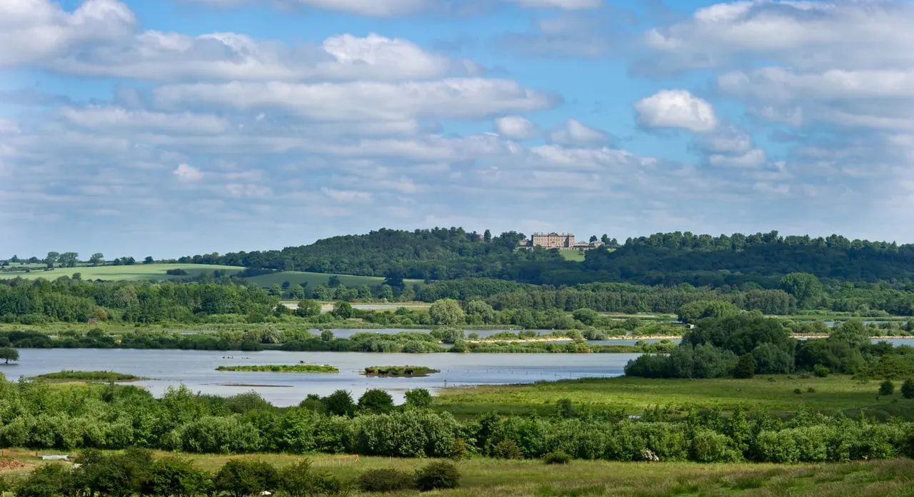 Expansive view of a lake with tree-covered islands, surrounded by green fields and woodland, with a stately home visible on the distant hillside.