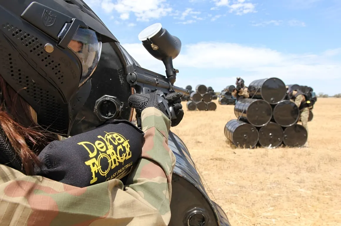 Player in camouflage holding paintball marker with barrel cover on sandy field, barrels stacked in background.