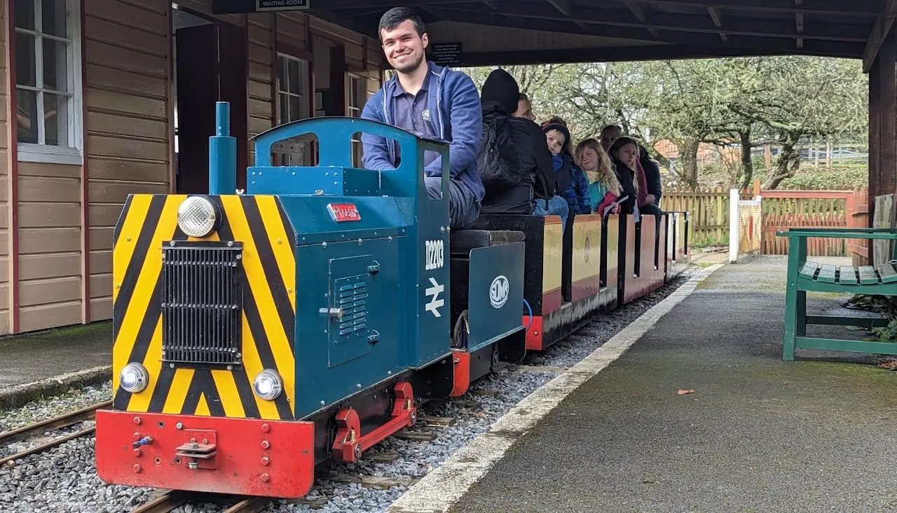 A man operates a blue and red miniature railway engine with yellow and black striped front whilst passengers sit in the attached carriages.