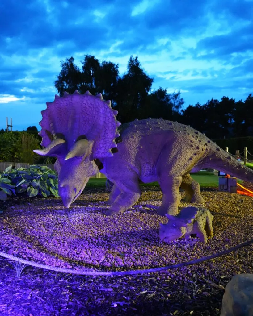 Large illuminated dinosaur sculpture with purple lighting in an outdoor adventure golf course at dusk.