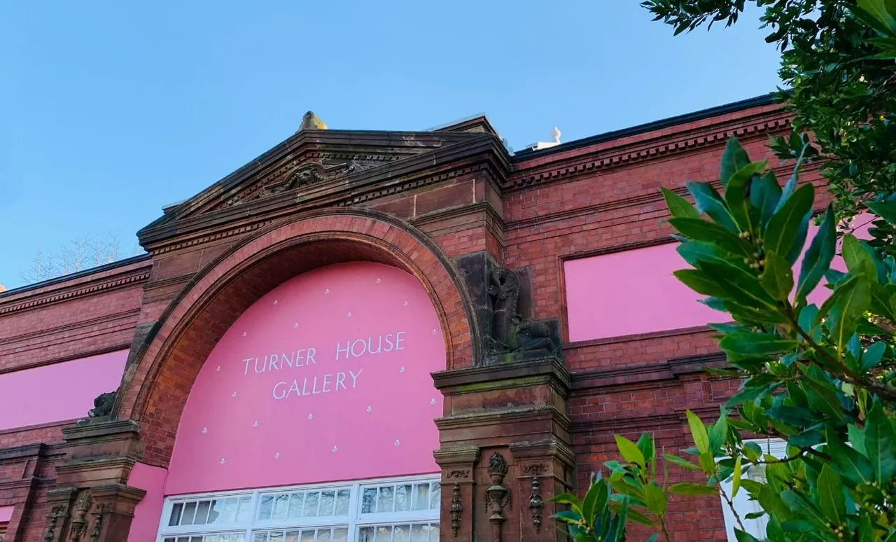 Victorian red-brick building with pink-painted archway entrance and signage for Turner House Gallery, framed by green foliage.