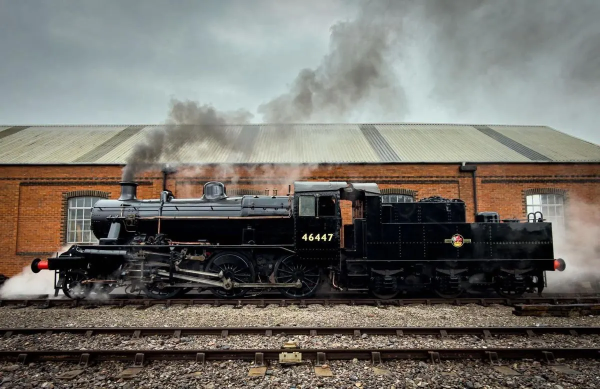 Steam locomotive number 46447 emitting smoke whilst stationary on tracks beside a brick railway building.