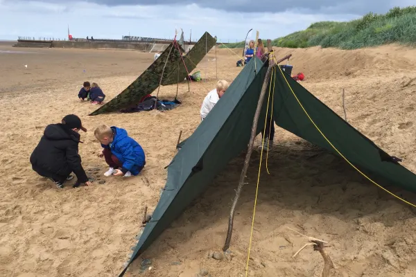 Children exploring tarps and camping shelters on a sandy beach with cliffs and sea wall visible.