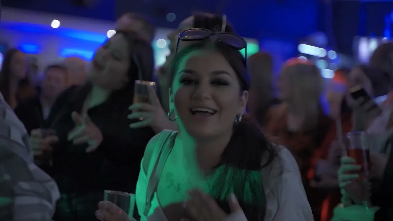 Woman smiling at a nightclub dance floor surrounded by other guests under blue and green lighting.